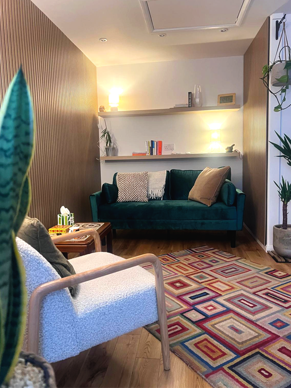 A picture of our counselling space. A light room, with a white seat facing away from, and a green sofa facing toward the camera. There is wooden slat panelling on the left and right walls, with shelves at the rear, behind the sofa. On the shelves are books and a couple of lamps, and on the floor, a patterned rug